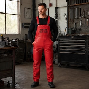 Op de afbeelding: Felrode leren tuinbroek met een borstzak met de zwarte tekst CUSTOM TEXT HERE, verstelbare schouderbanden en een rechte broekspijp. Het kledingstuk wordt gedragen over een zwart shirt met lange mouwen en zwarte schoenen in een werkplaats.