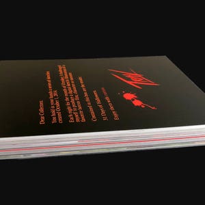 May include: A black book with red text and a red blood splatter design. The book is titled "31 Days of Halloween" and has the text "Enjoy them with" on the bottom. The book is stacked on top of other books.