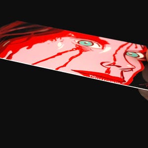 May include: A close-up of a woman's face with red paint splattered across her face and neck. Her eyes are wide open and her mouth is slightly open. The background is black.