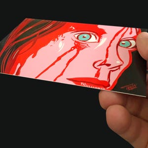 May include: A close-up of a woman's face with red paint dripping down her cheeks. Her eyes are wide open and her expression is one of fear. The background is black.