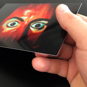May include: Close-up of a person's face with red and orange skin tones. The person has large, wide-set eyes with green irises. The image is painted in a stylized, almost abstract style.