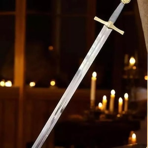 Puede incluir: Una espada de estilo medieval con una larga hoja plateada. La guarda es dorada y el pomo está decorado. La espada se muestra sobre un fondo oscuro, sugiriendo un tema histórico o de fantasía.