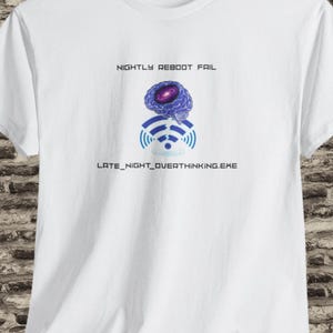 Op de afbeelding: Wit t-shirt met een grafisch ontwerp met een gestileerd brein, Wi-Fi-symbool en wolk. De tekst luidt "NIGHTLY REBOOT FAIL" en "LATE_NIGHT_OVERTHINKING.EXE". Het ontwerp is in blauw- en paars tinten.