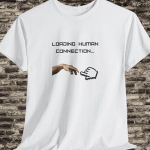 Op de afbeelding: Wit t-shirt met de tekst "LOADING HUMAN CONNECTION..." boven een illustratie van een hand die naar een pixelige computermuiscursor reikt. Het ontwerp is in zwart en wit.