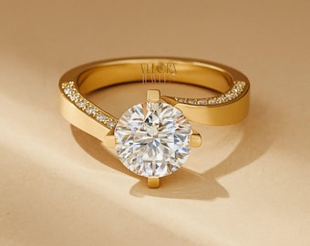 May include: A gold ring featuring a large, round, clear gemstone. The gold band is adorned with small, clear stones along the top. The ring has a modern, elegant design. The brand name "VELORA JEWEL" is visible on the ring.