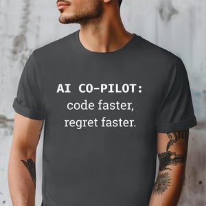 Op de afbeelding: Donkergrijs t-shirt met witte tekst: "AI CO-PILOT: code faster, regret faster." Het shirt met korte mouwen wordt door een persoon gedragen.