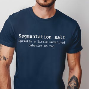Op de afbeelding: Marineblauw t-shirt met de witte tekst "Segmentation salt" en de zin "Sprinkle a little undefined behavior on top". Het shirt is effen van kleur en heeft korte mouwen.
