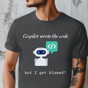 Op de afbeelding: Donkergrijs t-shirt met de tekst "Co-pilot wrote the code" boven een cartoonrobot en een tekstballon met codesymbolen. De tekst "but I get blamed?" staat eronder.