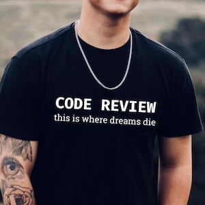 以下が含まれることがあります： 「CODE REVIEW」と「this is where dreams die」の文字が白でプリントされた黒いTシャツ。銀色のチェーンネックレスが首にかけられています。