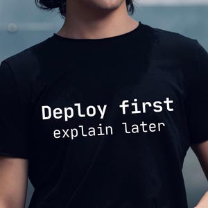 Könnte beinhalten: Schwarzes T-Shirt mit dem weißen Text "Deploy first explain later". Das Shirt hat einen Rundhalsausschnitt und kurze Ärmel. Der Text ist mittig auf der Vorderseite des Shirts platziert.