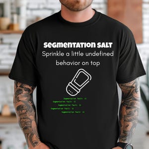 Op de afbeelding: Zwart T-shirt met de tekst "SEGMENTATION SALT Sprinkle a little undefined behavior on top." Een witte afbeelding van een zoutstrooier staat onder de tekst. De zin "Segmentation fault: 11" wordt in het groen herhaald.
