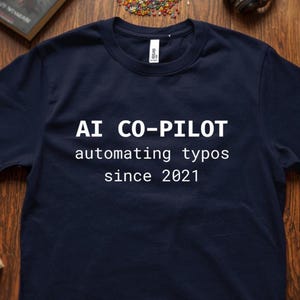 Könnte beinhalten: Marineblaues T-Shirt mit weißem Text "AI CO-PILOT automating typos since 2021". Das T-Shirt hat einen Rundhalsausschnitt und kurze Ärmel. Der Stoff scheint weich und bequem zu sein.