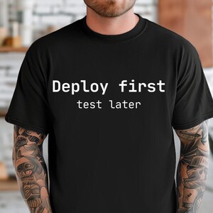Pode incluir: Camiseta preta de gola redonda com o texto branco "Deploy first test later". A camiseta é feita de um material macio e é adequada para uso casual. O texto está centralizado no peito.