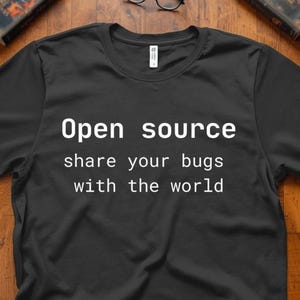 Può includere: T-shirt nera con la scritta bianca "Open source share your bugs with the world". La maglietta è a girocollo e sembra essere realizzata in un materiale morbido. La maglietta è esposta su una superficie di legno.