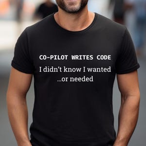 May include: Black t-shirt with white text: "CO-PILOT WRITES CODE, I didn't know I wanted...or needed." The shirt has a crew neck and short sleeves.
