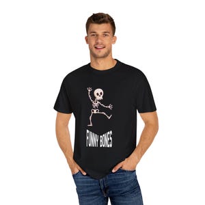 Può includere: T-shirt nera con un disegno di scheletro dei cartoni animati e la scritta "FUNNY BONES" in bianco. Lo scheletro è raffigurato in una posa giocosa. La maglietta è in tessuto morbido.