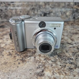 May include: Silver Canon PowerShot A80 digital camera with a Canon Zoom Lens 3x. The camera has a built-in flash and a lens that reads 7.8-23.4mm 1:2.8-4.9. The camera is on a speckled brown surface.