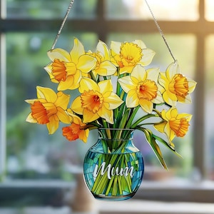 Op de afbeelding: Een gebrandschilderde raamdecoratie met een boeket gele en oranje narcissen in een blauwe vaas. De vaas heeft het woord "Mum" erop. Het raam hangt aan een zilveren ketting.