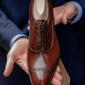 Peut inclure: Une chaussure de ville en cuir marron brillant avec une forme de chaussure en bois à l'intérieur. La chaussure présente un bout rapporté avec des perforations décoratives et des lacets marron foncé. La chaussure est tenue par une personne.