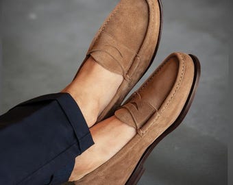 May include: A pair of light brown suede loafers with a classic penny strap design. The shoes have a dark brown sole and are stitched with a contrasting thread. The image shows the shoes from a low angle, highlighting their texture and shape.