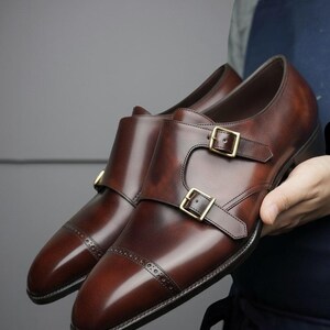 Pode incluir: Um par de sapatos de couro castanho escuro com fivela dupla, acabamento polido, biqueira com detalhes decorativos brogue e duas fivelas metálicas douradas em cada tira. Os sapatos têm uma silhueta elegante e uma sola escura.