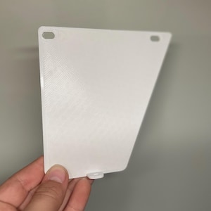 May include: A white, angled plastic component with a textured surface. The piece has two small, oval-shaped holes near the top and a small tab at the bottom. The item is held by a hand.