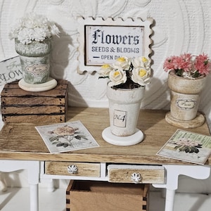 May include: Miniature wooden desk with white legs, holding small flower pots with white and pink flowers. A sign that says "Flowers Seeds & Blooms" is on the wall. Decorative cards and a wooden box are also on the desk.