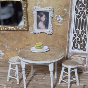 May include: Miniature dining set with a round table and two stools. The table has a light brown top and white legs. Two white plates and a yellow bowl sit on the table. A gold mirror and a framed portrait hang on the wall.