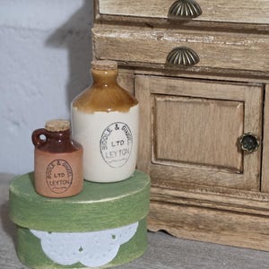 May include: Miniature ceramic bottles with cork stoppers, labeled "Biddle & Ginger LTD Leyton", sit on a green round box with a white lace design. A small wooden cabinet is in the background.
