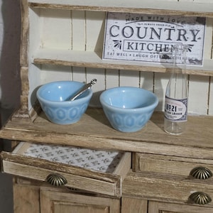May include: A miniature wooden hutch with a distressed finish, featuring two light blue bowls, a silver spoon, and a clear glass bottle with a label reading No 21 LES LEVRES. A sign above reads COUNTRY KITCHEN, and an open drawer shows a floral pattern.