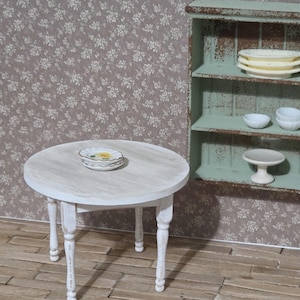 May include: A miniature white round table with four legs, topped with a stack of small plates. A distressed green shelf unit holds more plates and bowls. The background features a floral wallpaper, creating a vintage aesthetic.