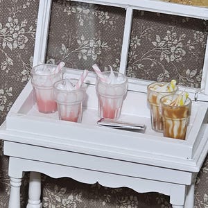 May include: Miniature white table with a tray holding several small clear glasses filled with pink and brown beverages. Some glasses have whipped cream and striped straws. The table has a white frame with a floral patterned background.
