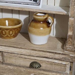 May include: A miniature ceramic jug with a glossy brown upper section and a white base, featuring a small handle. Beside it sits a matching brown ceramic bowl with a textured rim, both displayed on a wooden shelf with visible drawer pulls.