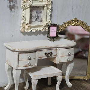 May include: A miniature white vanity set with a matching stool. The vanity has three drawers with crystal knobs and a small drawer with a bronze handle. A small framed picture and a gold-framed mirror are also present.