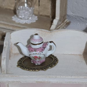 May include: A miniature porcelain teapot with pink floral designs and gold accents sits on a decorative brass-colored tray. The teapot is white with pink trim and a matching lid. The scene suggests a dollhouse or miniature tea set.