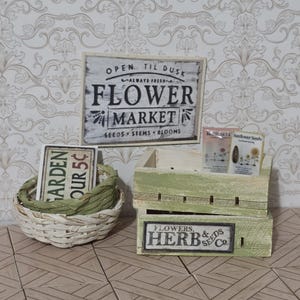 May include: A miniature flower market display featuring a rectangular sign reading OPEN TIL DUSK FLOWER MARKET, a small woven basket with green raffia, and a light green wooden crate labeled FLOWERS & HERB SEEDS CO with two seed packets.