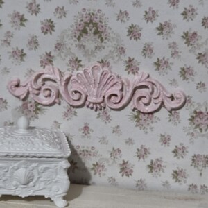 May include: A pale pink horizontal decorative molding featuring a central shell motif flanked by symmetrical scrolling acanthus leaf patterns, positioned above a white rectangular lidded box with intricate embossed floral detailing.