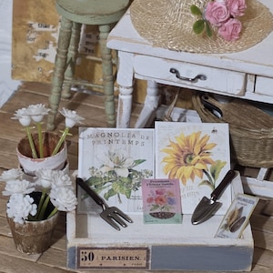 May include: A miniature garden scene featuring a white wooden tray labeled 50 PARISIEN, containing two seed packets, a metal trowel, and a metal fork. Nearby are two small pots with white flowers, a green stool, and a straw hat with pink roses.