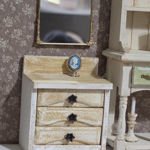 May include: Miniature bedroom set with a gold-framed mirror, a three-drawer nightstand, and a cameo figurine. The nightstand is painted white and beige with star-shaped drawer pulls. The mirror is ornate and hangs above the nightstand.