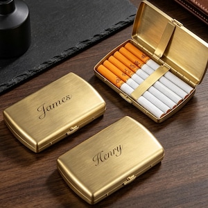 May include: Two rectangular brushed gold metal cigarette cases with rounded corners, one engraved with the name James and the other with Henry. A third open case displays six white cigarettes with orange filters held by a metal bar.