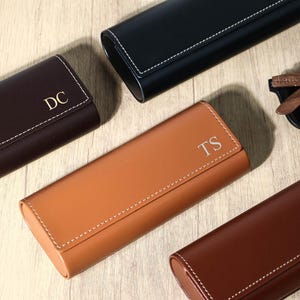 Könnte beinhalten: Eine Auswahl personalisierter Brillenetuis in Braun, Schwarz und Beige. Jedes Etui ist rechteckig mit abgerundeten Kanten und weißen Nähten. Einige Etuis haben goldfarbene Initialen. Eine Sonnenbrille ist ebenfalls zu sehen.