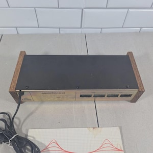 May include: An AudioControl electronic device featuring a black top panel, a gold-toned front face with printed technical specifications, and dark wood side panels. A black power cord with a two-prong plug extends from the left side of the unit.