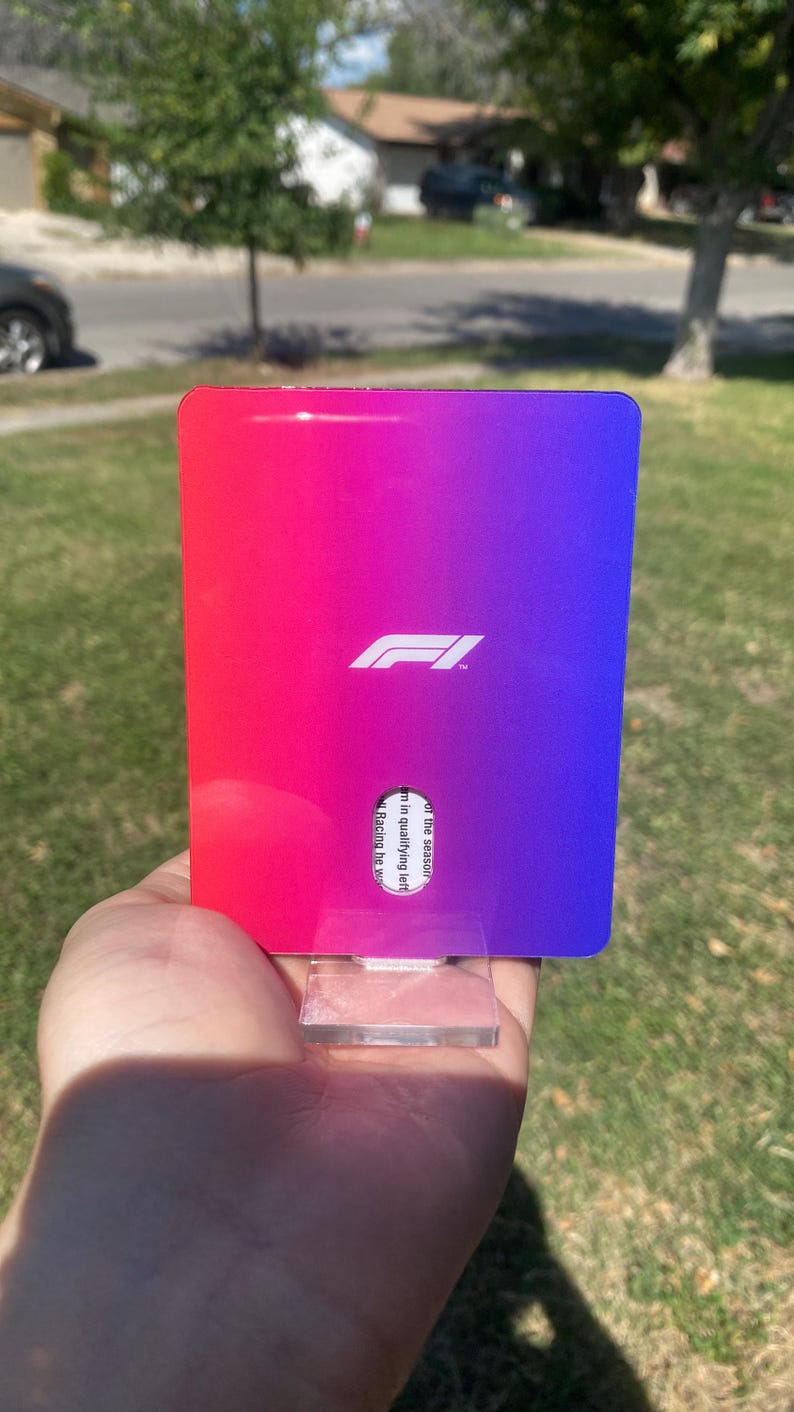 K&ouml;nnte beinhalten: Eine rechteckige Karte mit einem Farbverlauf von Rot nach Lila, mit dem wei&szlig;en Formula 1-Logo. Die Karte wird vor einem verschwommenen Hintergrund im Freien gehalten.
