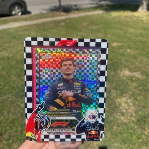 May include: A Max Verstappen Formula 1 trading card. The card features a photo of the driver in his Red Bull racing suit. The card has a checkered border and the F1 logo. The card is held in a clear plastic stand.