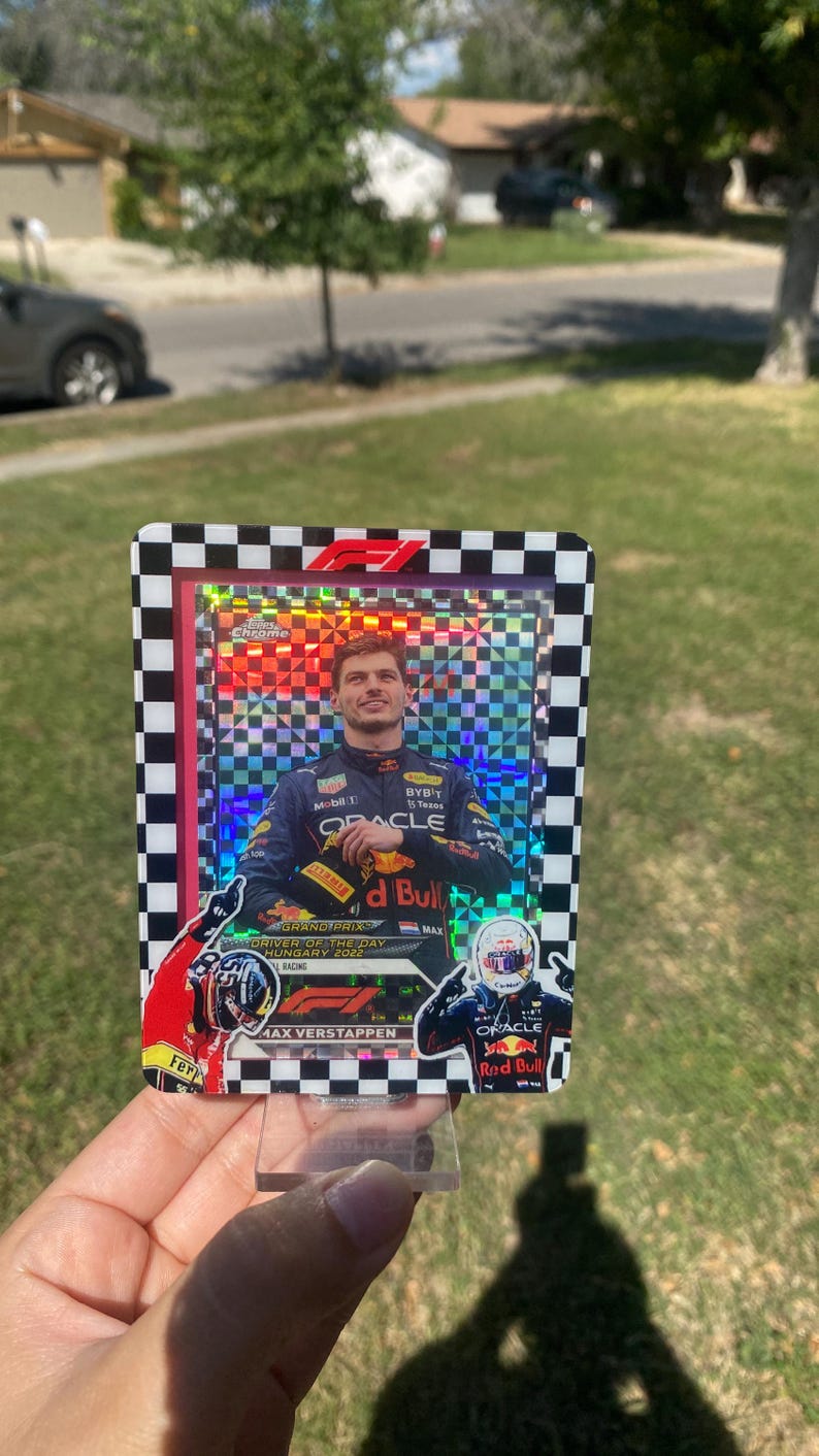 K&ouml;nnte beinhalten: Eine Max Verstappen Formel 1 Sammelkarte. Die Karte zeigt ein Foto des Fahrers in seinem Red Bull Rennanzug. Die Karte hat einen karierten Rand und das F1-Logo. Die Karte wird in einem transparenten Kunststoffst&auml;nder gehalten.