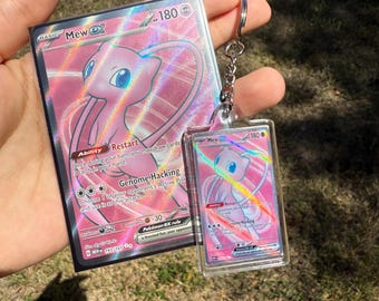 May include: A pink Mew EX Pokémon trading card and a matching keychain. The card features a pink Mew illustration with the text "Mew EX" and "180". The keychain has the same design, encased in a clear plastic holder.