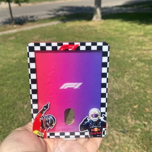 K&ouml;nnte beinhalten: Ein Formel-1-Kartenhalter mit einem Farbverlauf aus Rosa, Lila und Blau. Der Kartenhalter hat einen schwarz-wei&szlig; karierten Rand und zeigt das F1-Logo und Fahreraufkleber.