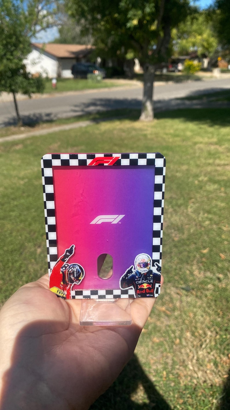 K&ouml;nnte beinhalten: Ein Formel-1-Kartenhalter mit einem Farbverlauf aus Rosa, Lila und Blau. Der Kartenhalter hat einen schwarz-wei&szlig; karierten Rand und zeigt das F1-Logo und Fahreraufkleber.