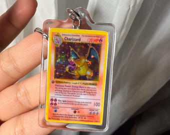 May include: A Charizard Pokémon card keychain. The card features a yellow and orange Charizard illustration with the text "Charizard" and "120 HP". The card is encased in a clear plastic holder with a silver-colored keyring.