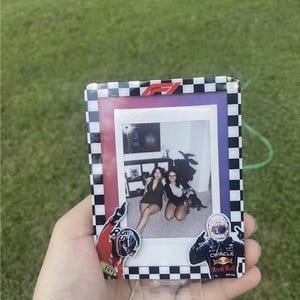 K&ouml;nnte beinhalten: Ein gerahmtes Foto mit einem schwarz-wei&szlig; karierten Rand, das ein Foto von zwei Personen zeigt. Der Rahmen hat einen roten und lila Innenrand und Formel-1-Logos. Aufkleber von Rennfahrern befinden sich auf dem Rahmen.